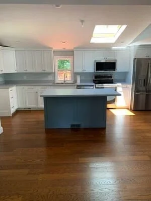 White painted kitchen cabinets with a slate blue painted island and hardwood floors under a skylight