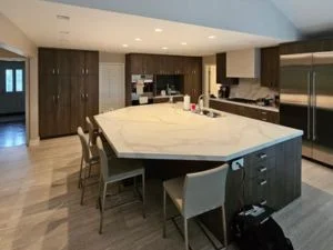 Kitchen with dark wood flat-panel cabinets, white veined countertop island, and gray upholstered stools on tile floor