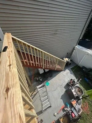 Unfinished pressure-treated wood staircase and railing beside vinyl siding, with concrete patio and work tools below