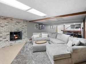 Finished basement living room with gray walls, dark wood ceiling beam, light stone fireplace, and gray sectional sofa